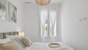 Een slaapkamer met een bed, witte gordijnen, een raam en een wandlamp.