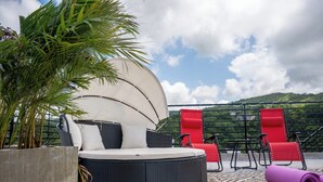 A rooftop lounge area with a round black wicker sofa, red and black lounge chairs, a large potted plant, and a rolled-up yoga mat on a patterned rug. - Rooftop Villa partial Sea view/ Mountain views (Ocho Rios)