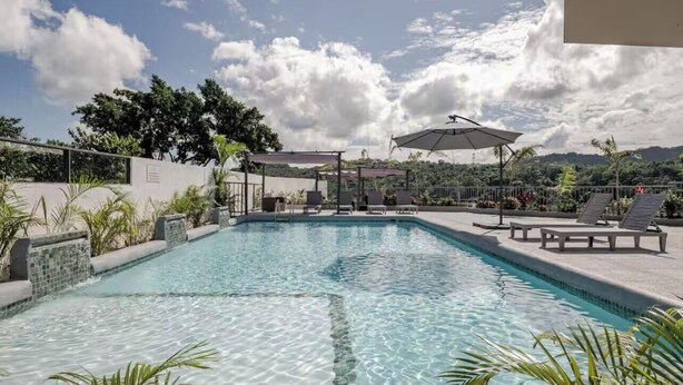 A rectangular swimming pool with clear water, surrounded by lounge chairs and umbrellas, set against a backdrop of trees and a cloudy sky. - Rooftop Villa partial Sea view/ Mountain views (Ocho Rios)