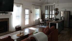 A spacious living room with a fireplace, a large television, and a ceiling fan. There is a dining area with a table and chairs, and a kitchen with a refrigerator and sink. - Beautiful, Cozy  and Airy Retreat with Lots of Character - NO CLEANING FEE (Clarksville)