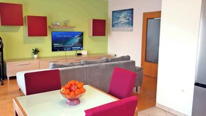 A modern living room with a flat-screen TV, grey sofas, a dining table with a bowl of fruit, and a green accent wall.