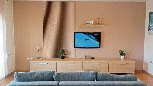 A modern living room with a flat-screen TV mounted on the wall, a wooden cabinet with drawers, a grey sofa, and a small potted plant.