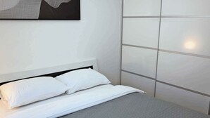 A neatly made bed with white bedding and a gray blanket, a white headboard, and a wall with a large, abstract artwork above it.