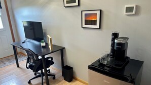Un pequeño espacio de oficina con un escritorio, una silla, una computadora, cuadros enmarcados, una máquina de café y un termostato.