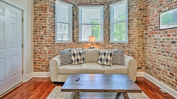A corner room with exposed brick walls, a beige sofa with patterned cushions, a wooden coffee table, and a window with blinds. - Charming Historic 1-bedroom apartment with WiFi, AC in pleasant Richmond (Richmond)