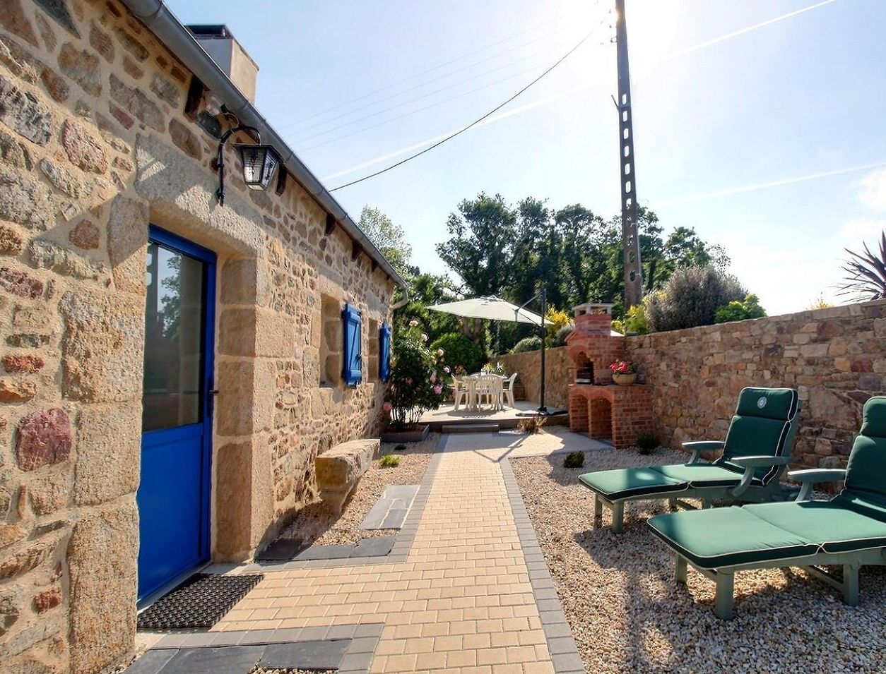 Un bâtiment en pierre avec une porte bleue, un espace extérieur aménagé avec des fauteuils de jardin verts et un chemin pavé menant à une terrasse.
