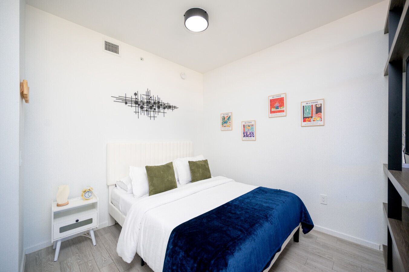 Quarto com cama branca, manta azul e mesa de cabeceira branca com abajur.