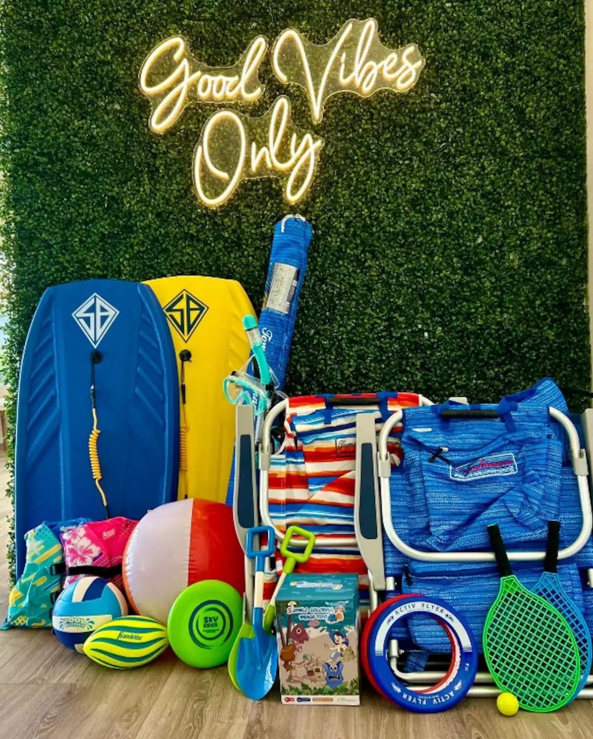 Varie attrezzature sportive e borse, tra cui una borsa blu e gialla, una borsa blu e a righe rosse e un racchetto da tennis blu e verde, sullo sfondo di un cartello con la scritta 'Good Vibes Only'.