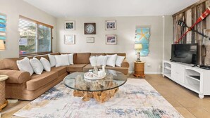 Un salón con un sofá modular, una mesa de centro de vidrio y un televisor sobre un mueble blanco.