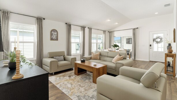 A spacious living room with a large sofa, a coffee table, and a TV. There are curtains on the windows, a plant on the side table, and a door with a wreath. - DISCOUNTED 2-bed Patio Villa in Waters Edge, near Eastport. Avail. Nov, Dec, Jan (The villages)