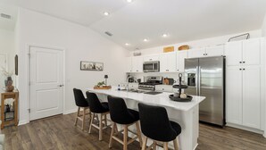 A modern kitchen with white cabinetry, a stainless steel refrigerator, a microwave, and a large island with bar stools. - DISCOUNTED 2-bed Patio Villa in Waters Edge, near Eastport. Avail. Nov, Dec, Jan (The villages)