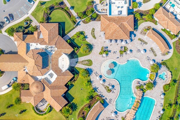 Vista aérea de un complejo turístico con una amplia piscina, varias zonas de descanso y un estacionamiento.