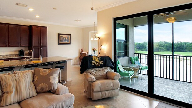 Un vaste espace de vie comprenant un canapé, un fauteuil inclinable et une table basse en verre. On y trouve également une cuisine avec des meubles en bois foncé et une table à manger. Un balcon avec vue sur l’extérieur est accessible par de grandes portes vitrées.