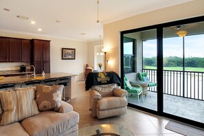 Amplia sala de estar con sofá, sillón reclinable y mesa de centro de vidrio. Se observa una cocina con armarios de madera oscura y una mesa de comedor. Se ve un balcón con vistas al exterior a través de amplias puertas de vidrio.