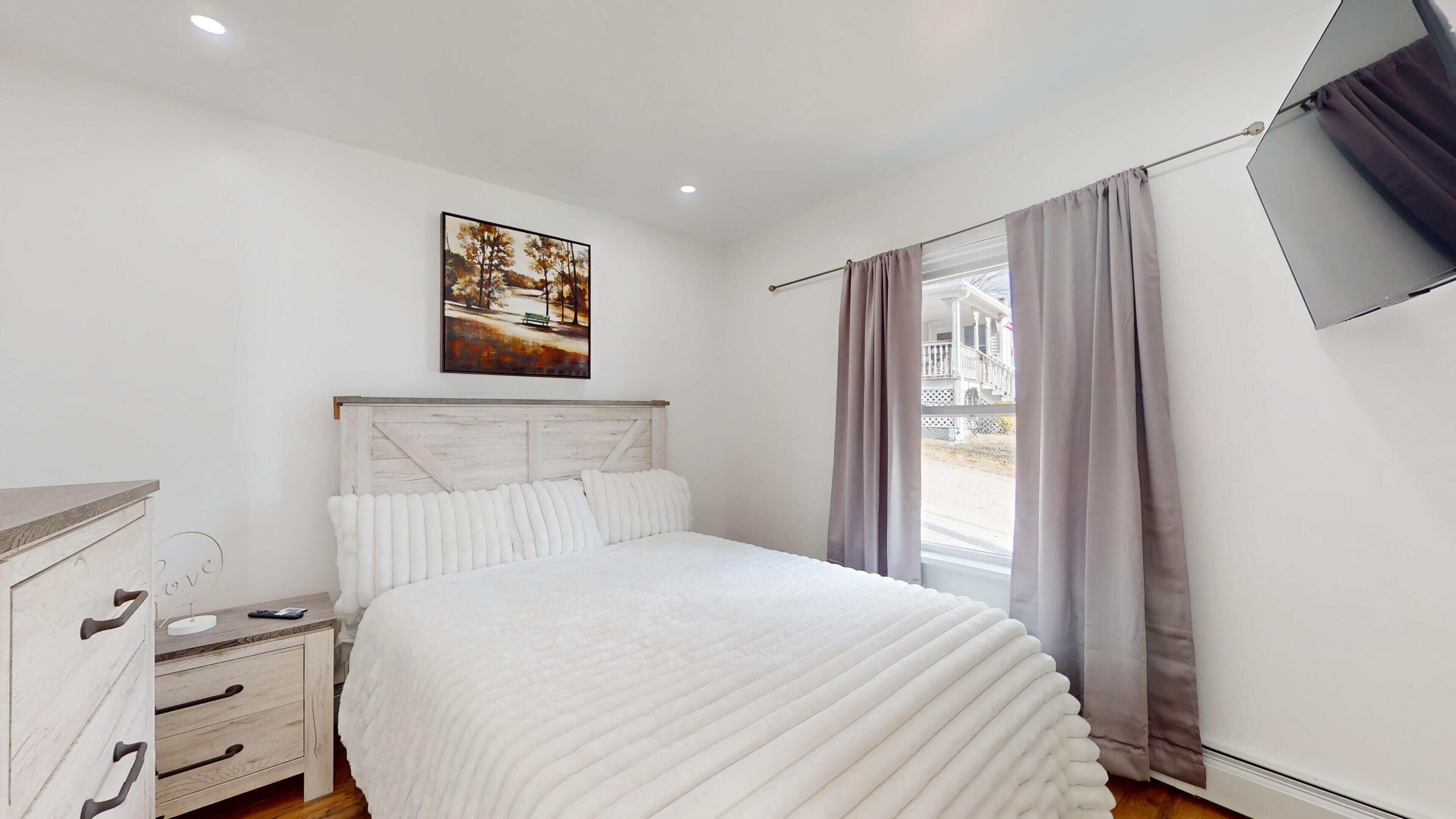 A bedroom with a bed, a nightstand, a dresser, a painting on the wall, and a window with curtains.