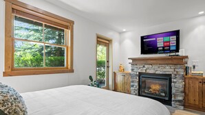 A bedroom with a large bed, a stone fireplace, a television mounted above it, and a window with a view of greenery.