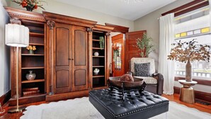 A spacious living room with a large wooden cabinet, a black leather ottoman, a white rug, and a view of the city through a large window.