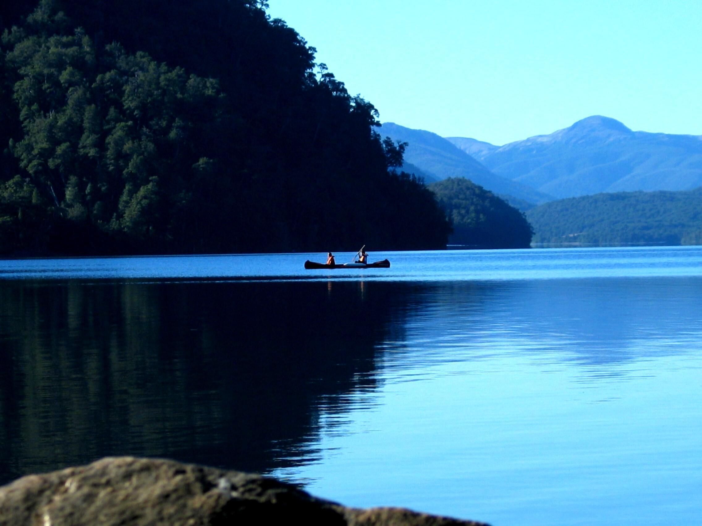 A calm lake with a canoe in the center, surrounded by forested hills and mountains in the background.