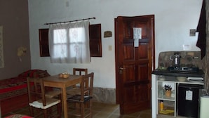 Une petite pièce chaleureuse, avec une table et des chaises en bois, un lit recouvert d’une couverture à motifs, une porte en bois et une kitchenette équipée d’une cuisinière et d’étagères.
