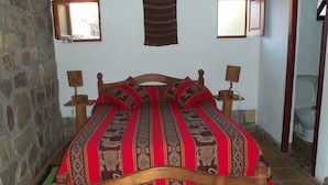 Un lit avec un cadre en bois et une couvre-lit à motifs rouges et noirs. Il y a deux petites fenêtres sur le mur, et un mur de pierre est visible sur la gauche.
