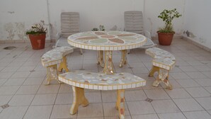 Een ronde buitentafel met mozaïektegels als zitbankjes, omringd door potplanten en twee comfortabele stoelen.