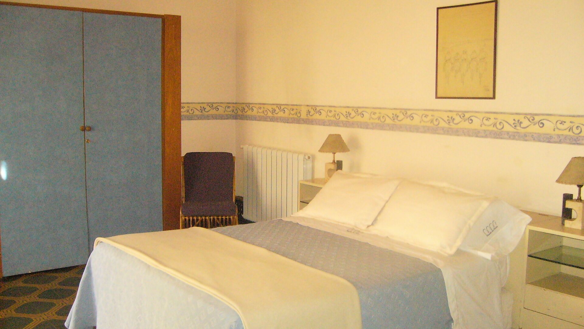 A hotel room with a bed, two bedside lamps, a chair, a radiator, and a wardrobe.