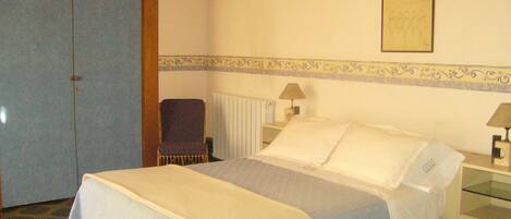 A hotel room with a bed, two bedside lamps, a chair, a radiator, and a wardrobe.
