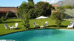 Una piscina con acqua cristallina circondata da un prato verde e alberi. Sono presenti sdraio bianche e un ombrellone che offre ombra.