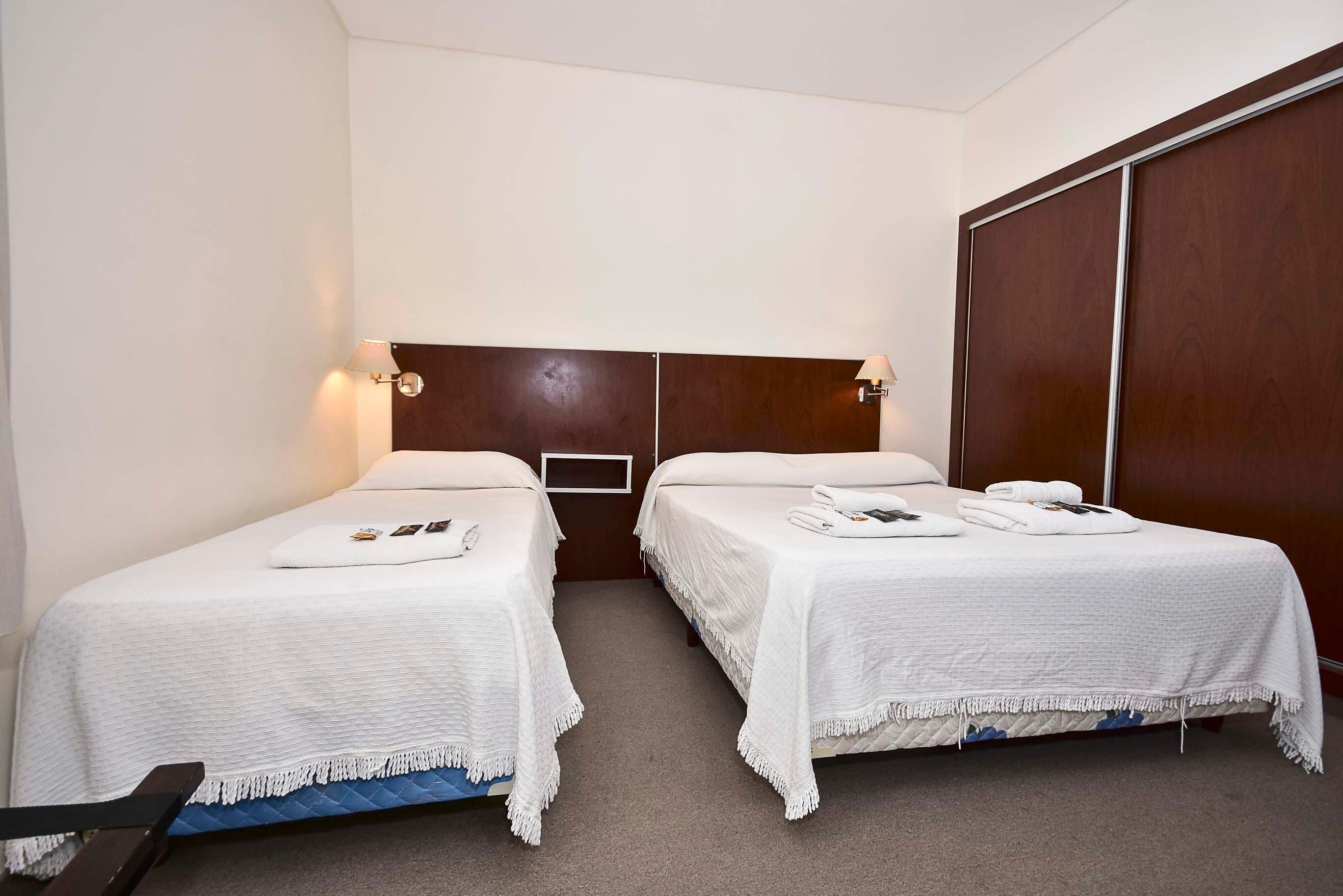 A hotel room with two single beds, a wooden headboard, a wardrobe, and a small table with a chair.