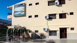 A hotel with a large sign advertising the Gran Hotel Tomasso di Savoia, featuring a coastal scene. There are outdoor seating areas with chairs and tables, and the building has multiple air conditioning units. - Gran Hotel Tomaso Di Savoia (La Falda)