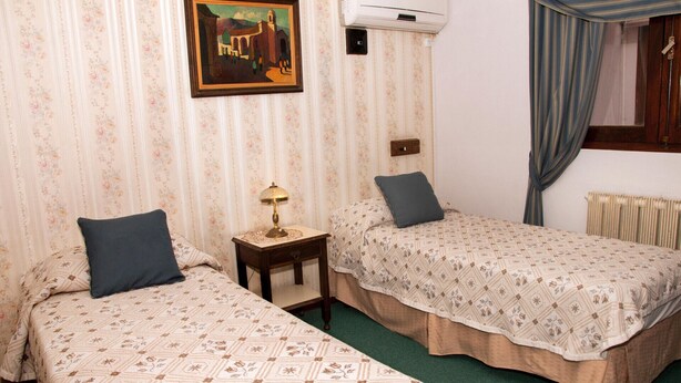 A room with two beds, floral-patterned bedding, a small table with a lamp, a painting on the wall, and an air conditioning unit. - Gran Hotel Tomaso Di Savoia (La Falda)