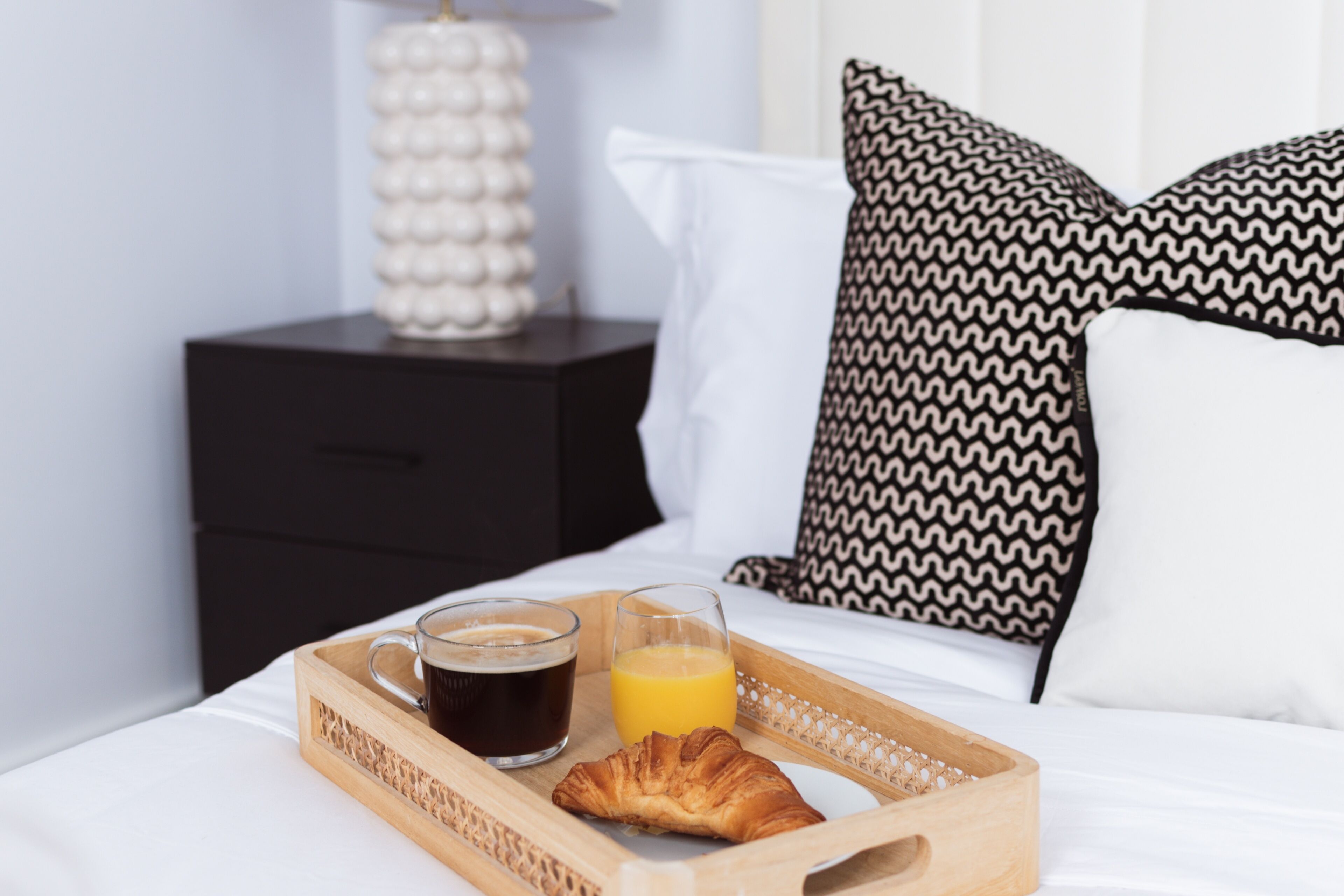 Un plateau de petit-déjeuner avec du café, du jus d’orange et un croissant, posé sur un lit avec une literie blanche et des oreillers à motifs.