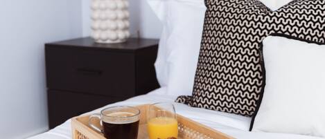 Ein Frühstücksbrett mit Kaffee, Orangensaft und Croissant auf einem Bett mit weißer Bettwäsche und gemusterten Kissen.