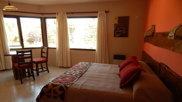 Une chambre d’hôtel avec un lit, un bureau et des chaises. Il y a des fenêtres avec des rideaux, un tableau au mur et un sol carrelé.