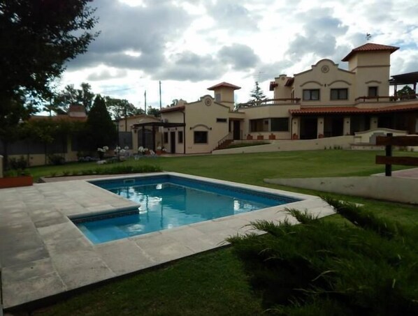 A large, rectangular swimming pool surrounded by a paved deck, with a well-maintained lawn and a multi-story building in the background.