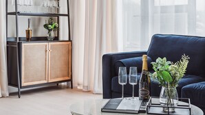 A modern living room with a dark blue sofa, a glass coffee table, a bookshelf with wine glasses, and a bottle of champagne. - Stylish new-build FREE parking, 10min to BHX & NEC (Birmingham)