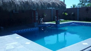 A swimming pool with a thatched roof structure nearby, surrounded by a brick wall and a small garden area. - Hotel Rincón Mágico (El Fuerte)
