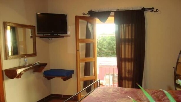A bedroom with a bed, a television, a window with curtains, and a small shelf. - Hotel Rincón Mágico (El Fuerte)