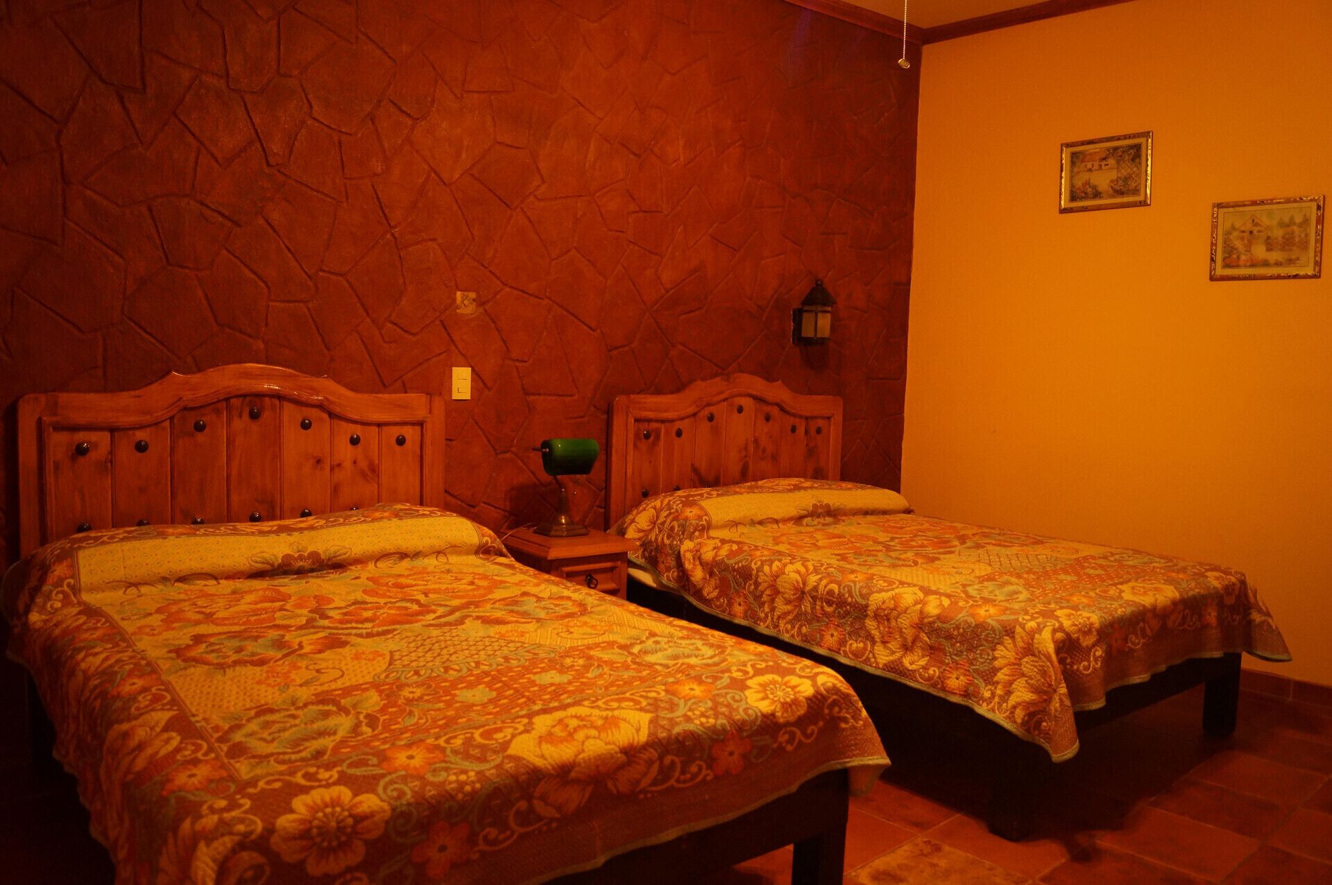 Ein Zimmer mit zwei hölzernen Betten, die mit blumigen Überwürfen ausgestattet sind, einer Steinwand und einem kleinen Tisch mit einer Lampe.
