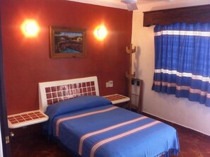 A room with a bed, a small table, a chair, a painting on the wall, and a window with curtains. - Mision Mares (Santa María Huatulco)