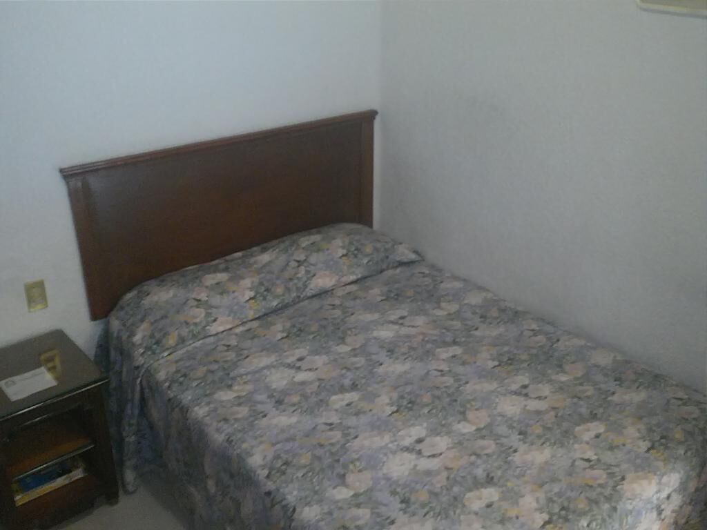 Habitació petita amb un llit individual, capçalera de fusta, cobrellit floral i una tauleta de nit amb una làmpada.