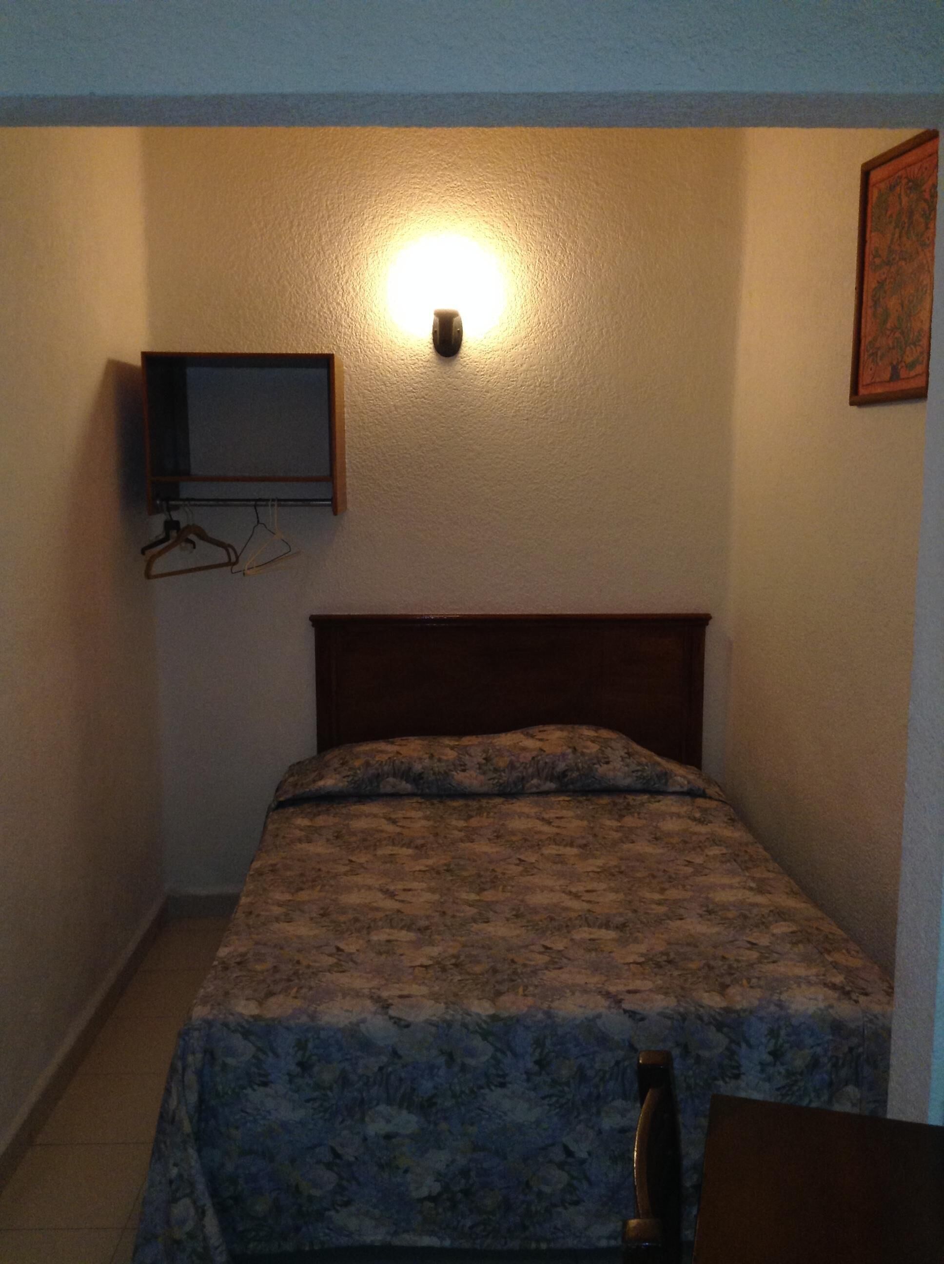 Een kleine, zwak verlichte kamer met een enkel bed, een aan de muur bevestigde televisie en een lijst met een afbeelding.