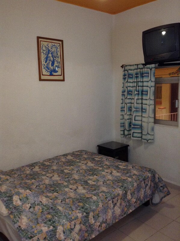 A bedroom with a floral bedspread, a framed picture on the wall, a small bedside table, and a television on a stand. - Hotel America (Cuernavaca)