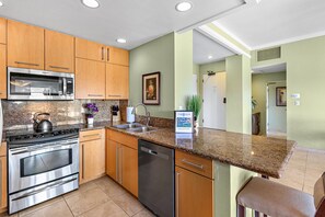 A modern kitchen with stainless steel appliances, wooden cabinets, granite countertops, and a view of the living area.