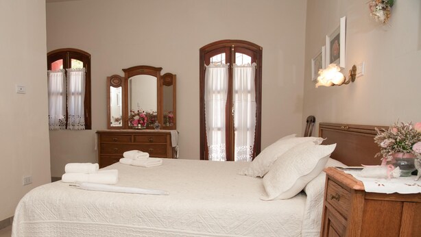 Een net opgemaakt bed met witte lakens, een houten dressoir en een spiegel met een bloemstuk.