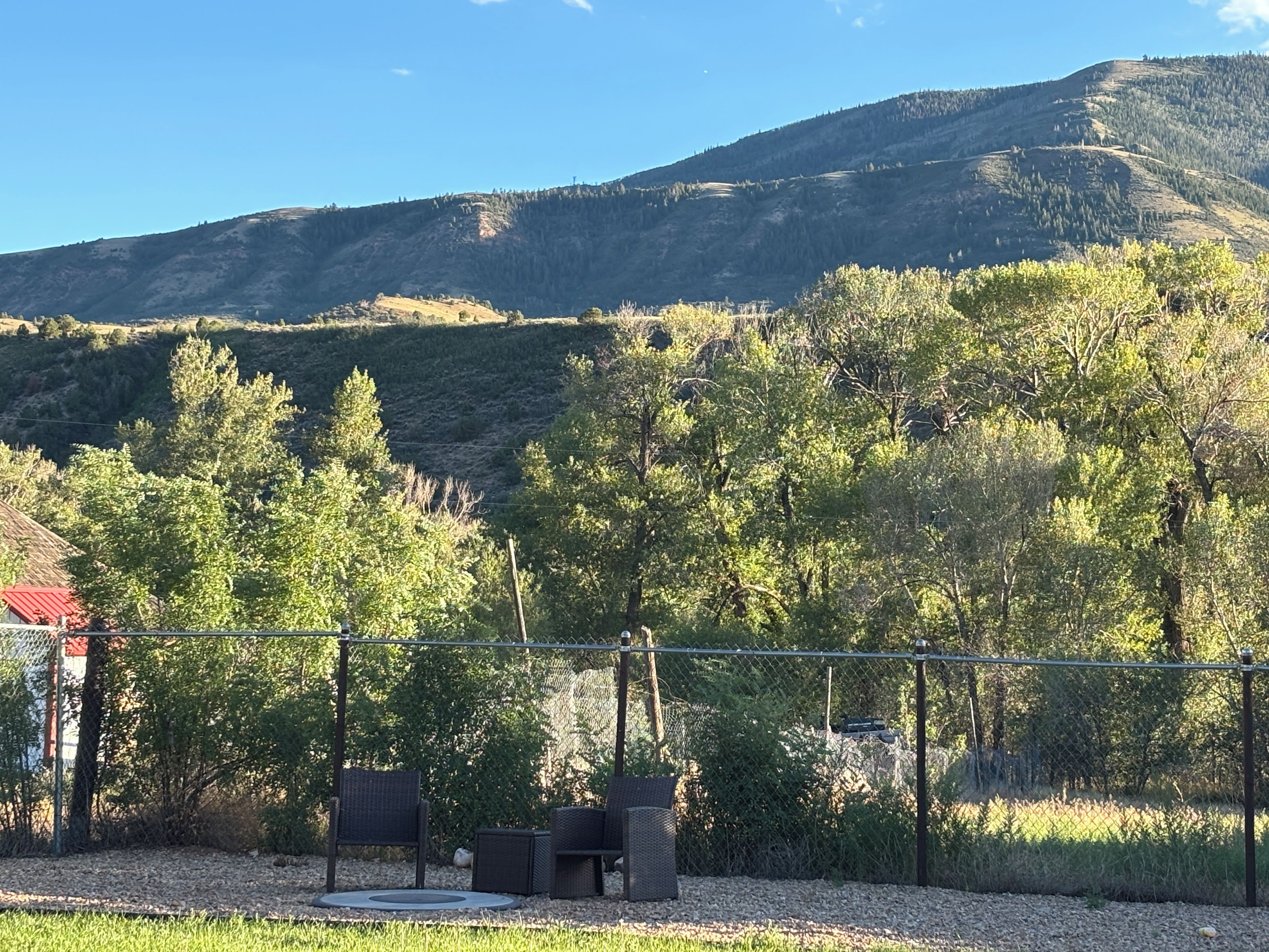 A fenced outdoor area with a table and chairs, surrounded by trees and a mountain in the background.