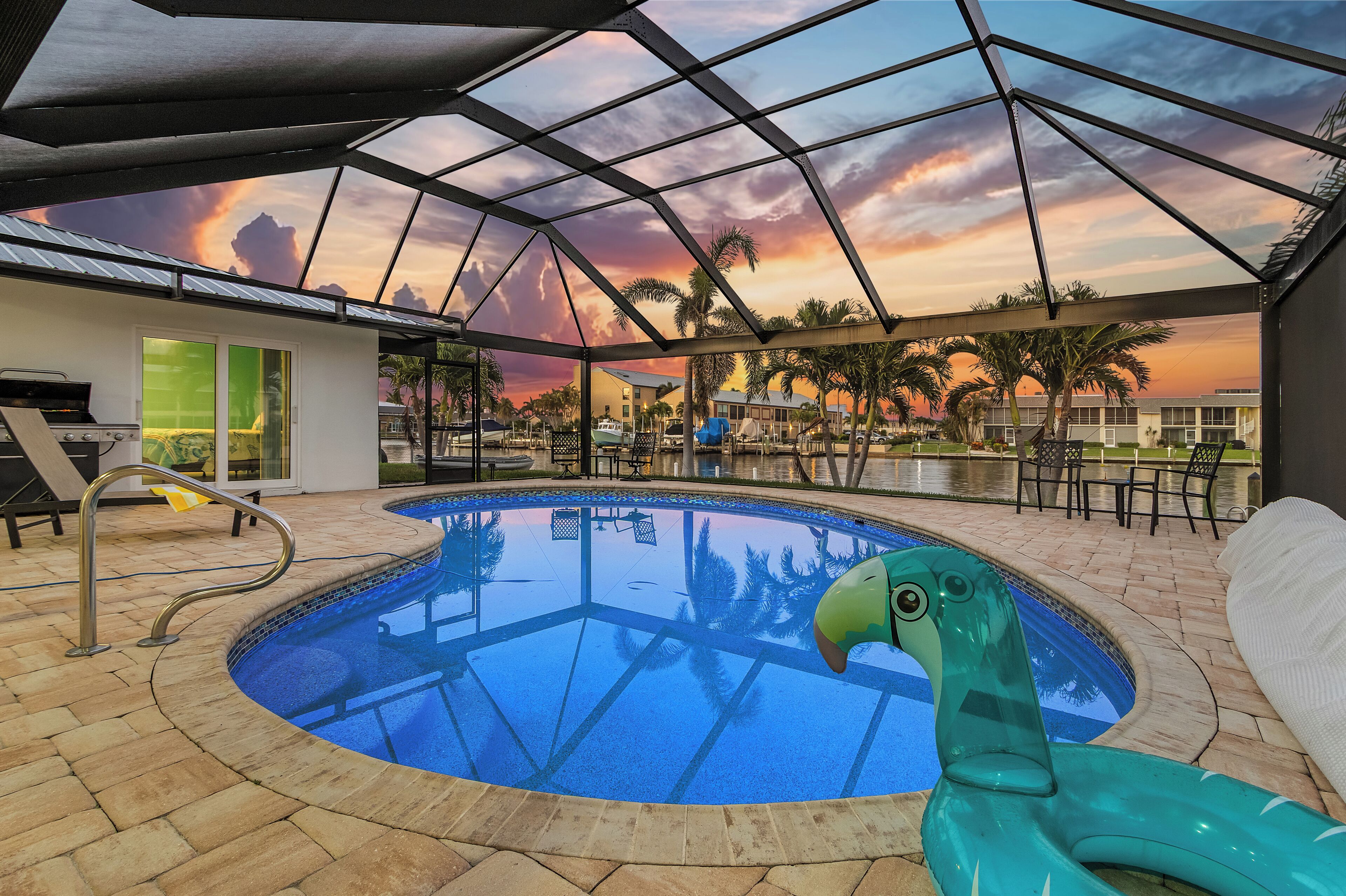 En indendørs swimmingpool med et glasloft, en grøn flamingo-flyder og udsigt til palmer og bygninger.