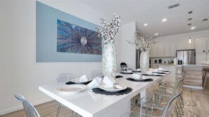 A modern dining area with a long table set for six, clear chairs, and decorative vases. - 12BR Solterra Mansion + Arcade Game Room & Pool (Davenport)