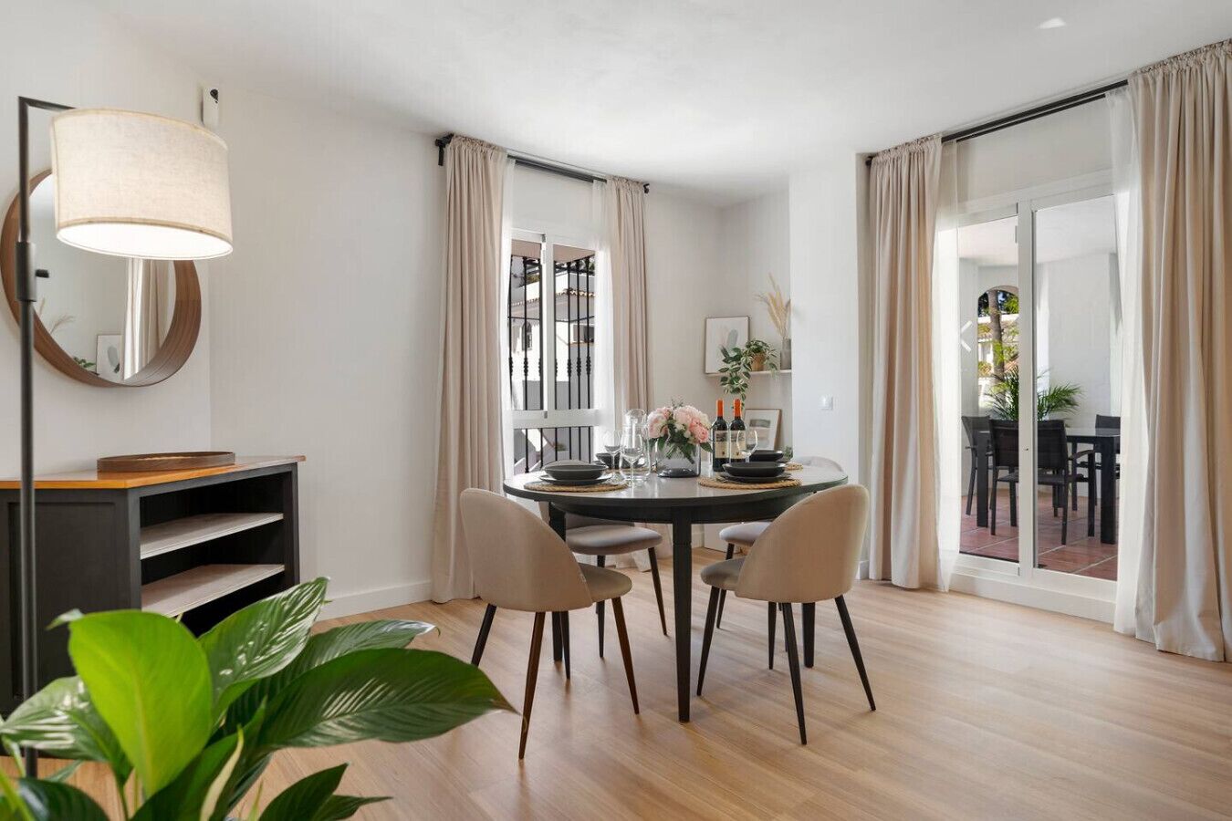 Ein Essbereich mit einem runden Tisch, Stühlen und einer Essgruppe. Im Vordergrund steht eine Pflanze und durch das Fenster ist eine Terrasse im Freien zu sehen.