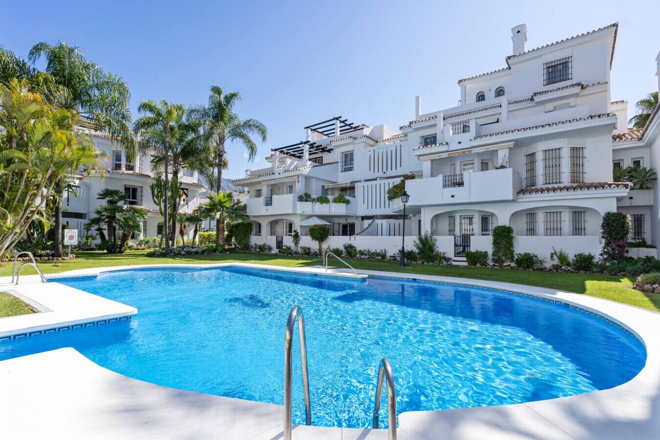 Una gran piscina rodeada de edificios blancos con balcones, palmeras y un cielo despejado.